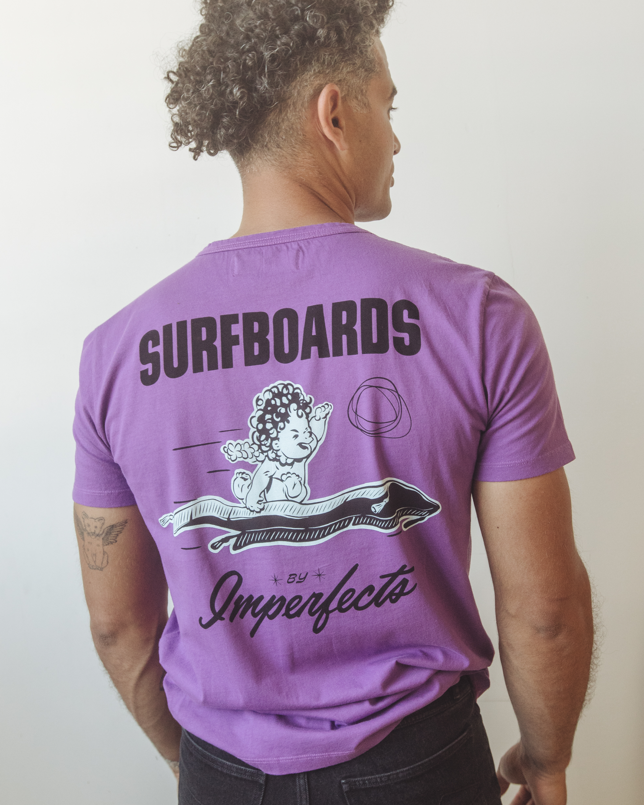 Back of men’s purple organic cotton t-shirt with bold “Surfboards by Imperfects” text and a hand-drawn cherub riding a surfboard, representing the iconic Magic Carpet model.
Magic Carpet T-Shirt in Purple Magic.
