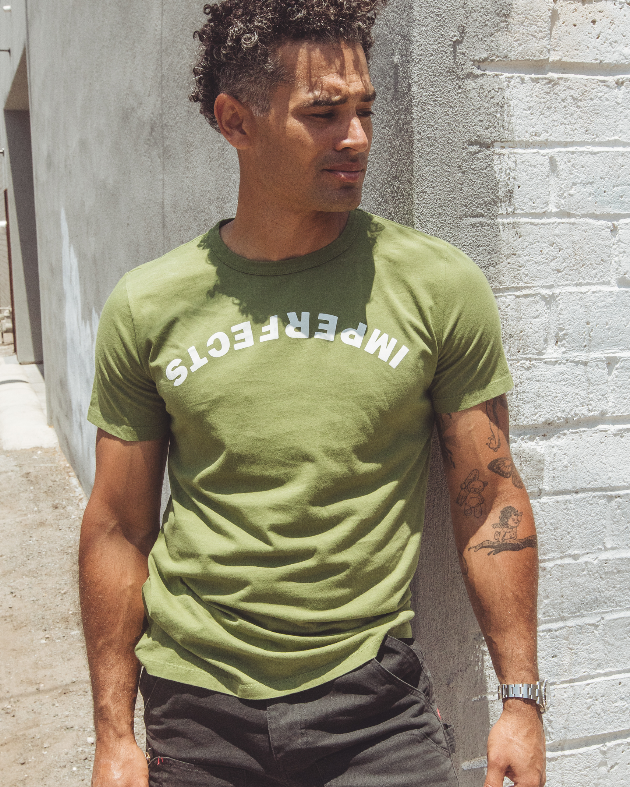 Men’s fatigue green t-shirt made from washed organic cotton, featuring bold arched “IMPERFECTS” logo flipped upside down across the chest, styled with dark trousers.
Upside Down T-Shirt in Fatigue.