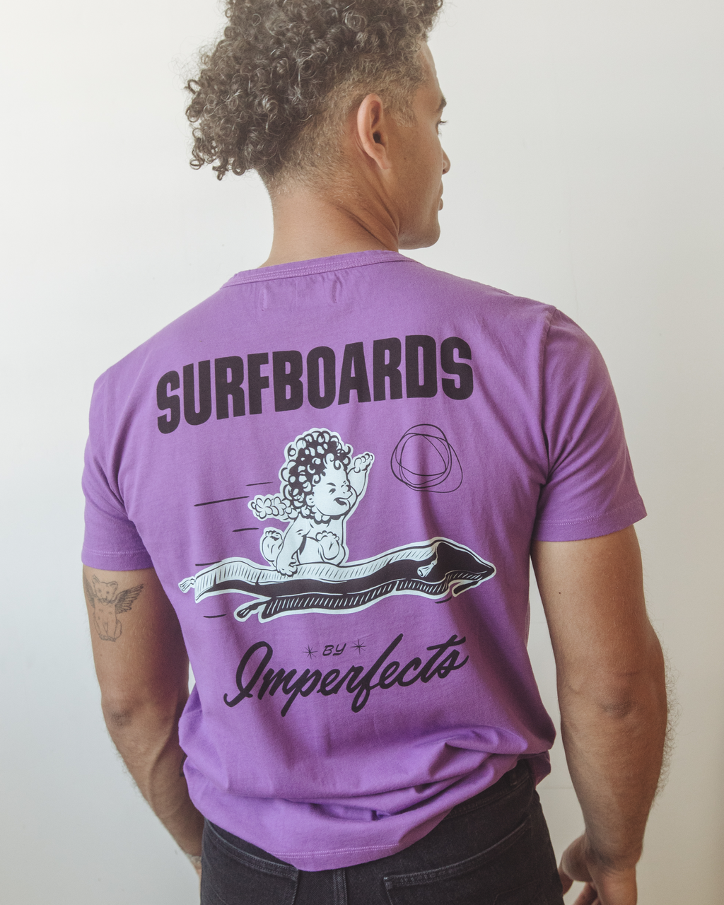 Back of men’s purple organic cotton t-shirt with bold “Surfboards by Imperfects” text and a hand-drawn cherub riding a surfboard, representing the iconic Magic Carpet model.
Magic Carpet T-Shirt in Purple Magic.