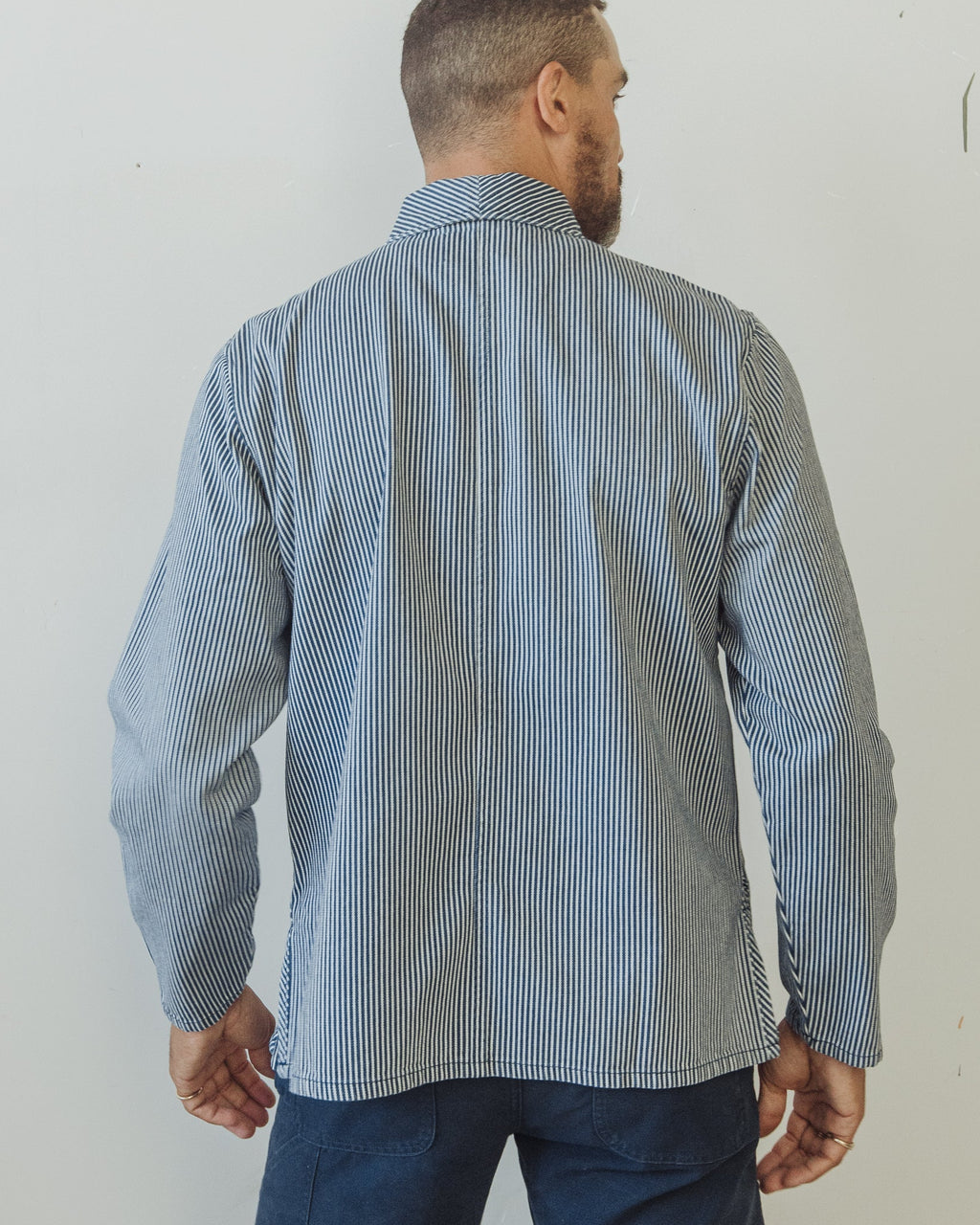 Man wearing a blue striped shirt and dark pants against a white wall. Shepherds Shirt in Indigo Hickory Stripe Original. O.G.
